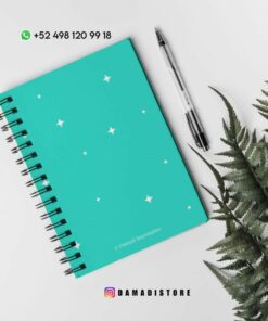 Un cuaderno de espiral verde azulado con estrellas blancas en la portada se coloca sobre una superficie blanca junto a un bolígrafo negro. Las hojas de helecho están situadas a la derecha. En la parte superior hay un número de WhatsApp y en la parte inferior hay un identificador de Instagram.