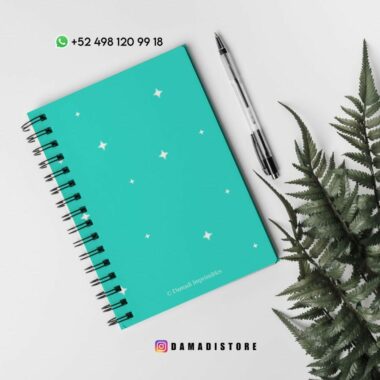 Un cuaderno de espiral verde azulado con estrellas blancas en la portada se coloca sobre una superficie blanca junto a un bolígrafo negro. Las hojas de helecho están situadas a la derecha. En la parte superior hay un número de WhatsApp y en la parte inferior hay un identificador de Instagram.