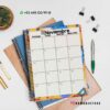 Un calendario de noviembre de 2023 se encuentra encima de cuadernos de espiral junto con varios suministros de oficina, incluidos un bolígrafo, un lápiz, una planta, sujetapapeles y clips. El calendario presenta un borde colorido y detalles de contacto, incluido un número de teléfono y identificadores de redes sociales.