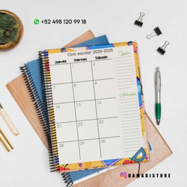 Sobre un escritorio hay una agenda abierta titulada "Ciclo escolar 2024-2025", que muestra los bloques del calendario de los jueves, viernes y sábados. Cerca hay cuadernos, un bolígrafo, clips para carpetas, una pequeña planta en una maceta y datos de contacto. La agenda lleva la marca "Damadistore".