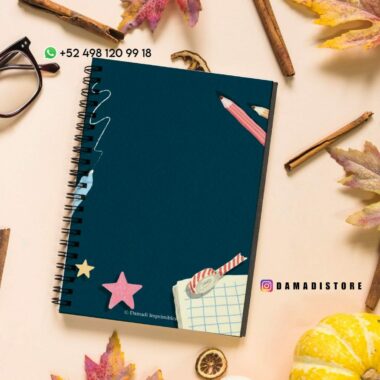 Sobre una mesa hay un cuaderno de espiral con tapa azul oscuro, rodeado de hojas de otoño, un par de vasos y una pequeña calabaza. La portada presenta recortes coloridos de estrellas, un clip, un lápiz y una hoja de papel cuadriculado. Los datos de contacto están en la esquina.