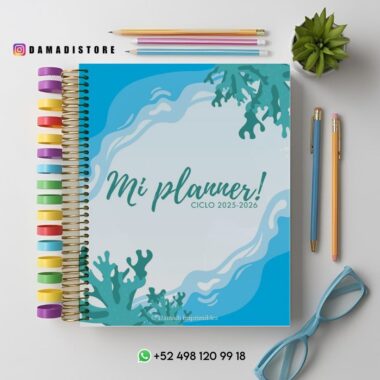 Una agenda titulada "¡Mi agenda! CICLO 2025-2026" con una portada azul océano con diseños coralinos. Pestañas de colores en el lado izquierdo. Cerca hay unas gafas, una pequeña planta verde y la información de contacto de Damadistore en la parte inferior.