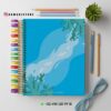 Un colorido cuaderno de espiral con una cubierta azul con temática oceánica y algas. Alrededor hay lápices de colores, gafas azules, una planta verde y la información de contacto de una tienda.