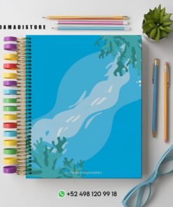 Un colorido cuaderno de espiral con una cubierta azul con temática oceánica y algas. Alrededor hay lápices de colores, gafas azules, una planta verde y la información de contacto de una tienda.