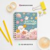 Una agenda colorida para profesor, con estrellas, nubes y un planeta en la portada, reposa sobre un escritorio blanco con lápices amarillos, clips, cinta adhesiva y una taza de café. También se ven los datos de contacto de Instagram y WhatsApp.