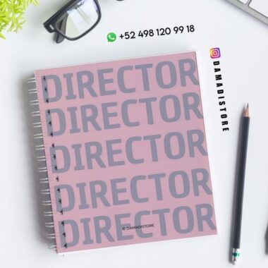 Un cuaderno de espiral rosa con la palabra "DIRECTOR" repetida en grandes letras grises en la portada, colocado sobre un escritorio blanco con un lápiz, una planta, gafas e información de contacto de DAMADISTORE.