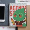 Un cuaderno encuadernado en espiral con un simpático personaje verde de dibujos animados que sostiene un lápiz y una regla en la portada, junto a un smartphone y un papel. En la imagen aparece "DAMADISTORE" y la información de contacto.