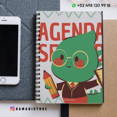 Un cuaderno encuadernado en espiral con un simpático personaje verde de dibujos animados que sostiene un lápiz y una regla en la portada, junto a un smartphone y un papel. En la imagen aparece "DAMADISTORE" y la información de contacto.