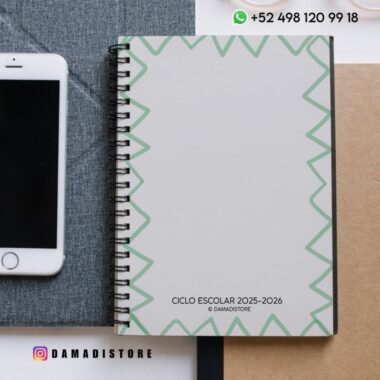 Un cuaderno encuadernado en espiral con un borde verde geométrico en la portada con la etiqueta "Ciclo Escolar 2025-2026", colocado junto a un smartphone. Se ve el contacto de WhatsApp y la cuenta de Instagram de DAMADISTORE.