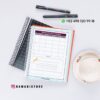 Agenda docente primaria 2025-2026 18 Colocación plana de cuadernos, bolígrafos y una taza de té sobre una superficie blanca. El cuaderno abierto muestra una página de la agenda semanal en español. Se ven los datos de contacto de WhatsApp y la cuenta de Instagram @damadistore.