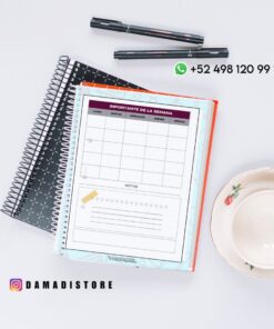 Colocación plana de cuadernos, bolígrafos y una taza de té sobre una superficie blanca. El cuaderno abierto muestra una página de la agenda semanal en español. Se ven los datos de contacto de WhatsApp y la cuenta de Instagram @damadistore.