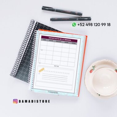 Colocación plana de cuadernos, bolígrafos y una taza de té sobre una superficie blanca. El cuaderno abierto muestra una página de la agenda semanal en español. Se ven los datos de contacto de WhatsApp y la cuenta de Instagram @damadistore.