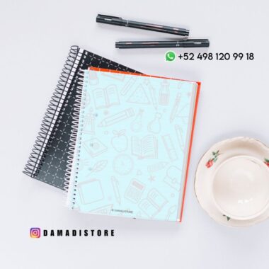 Tres cuadernos de espiral, dos bolígrafos y una taza de té con platillo sobre una superficie blanca. El cuaderno superior tiene una cubierta azul claro con ilustraciones de temática escolar. El texto de la imagen incluye un número de teléfono y una cuenta de Instagram.