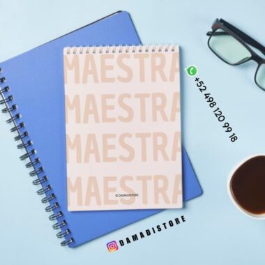 Encima de un cuaderno azul hay un bloc de notas en espiral con la palabra "MAESTRA" repetida en letras grandes de color rosa claro. Cerca hay un par de gafas, una taza de café e información de contacto de DAMADISTORE.