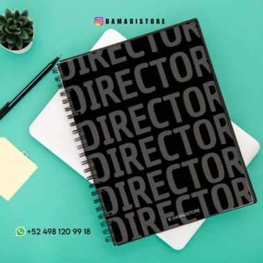 Un cuaderno negro de espiral con la palabra "DIRECTOR" repetida en negrita descansa sobre una tableta blanca. Cerca hay un bolígrafo, notas adhesivas y una pequeña maceta. En la imagen aparecen los datos de contacto y la cuenta de Instagram @damadistore.