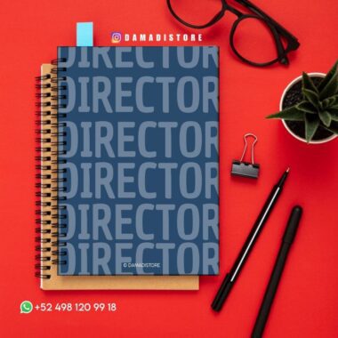 Un cuaderno azul con la palabra "DIRECTOR" impresa repetidamente en la portada está colocado encima de un cuaderno marrón con espiral. Alrededor hay unas gafas negras, una planta, un clip para carpetas, bolígrafos negros y un número de contacto de WhatsApp sobre una superficie roja.