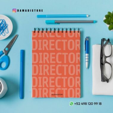 Un cuaderno de espiral con "DIRECTOR" repetido en negrita en la portada reposa sobre un escritorio azul rodeado de un bolígrafo, un lápiz, gafas, tijeras, clips de carpeta, clips de papel y una pequeña planta. Se ven los contactos de Instagram y WhatsApp.