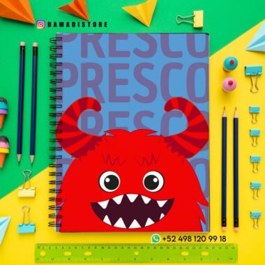 Un cuaderno de espiral con un monstruo rojo de dibujos animados y grandes dientes en la portada, rodeado de lápices de colores, clips, papel y una regla amarilla sobre un fondo azul y amarillo. La información de contacto de Damadistore es visible.