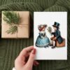 Niños Victorianos Clipart Bundle. Png, Svg, Eps, Jpg, Pdf. 3 Una mano sostiene una tarjeta de estilo antiguo que representa a un niño con sombrero de copa que ofrece flores a una niña con cofia. La tarjeta está sobre una superficie tejida de color verde, junto a un regalo envuelto y adornado con una ramita de vegetación.